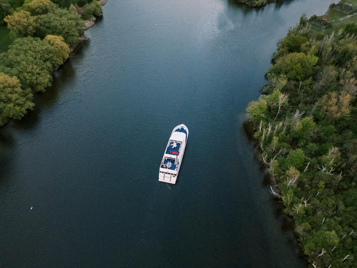 Aerial view of a white cruise ship sailing through a narrow, tree-lined river with lush greenery on both sides, under a clear blue sky.