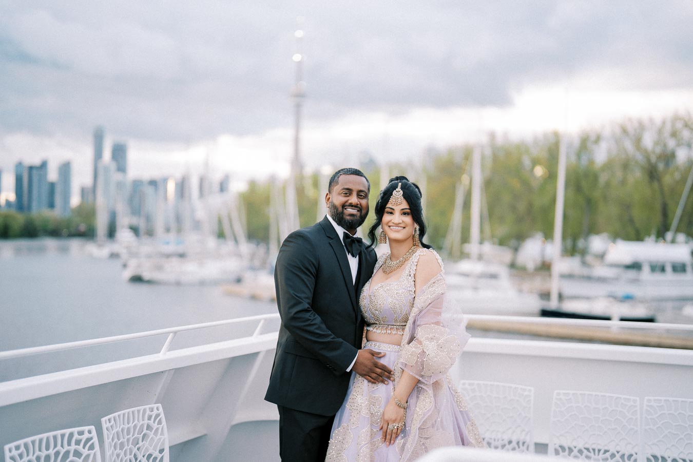 A couple in formal attire, standing on a boat deck with a city skyline and marina in the background. The woman is wearing an intricately designed lavender outfit with traditional jewelry, while the man is dressed in a classic black tuxedo.