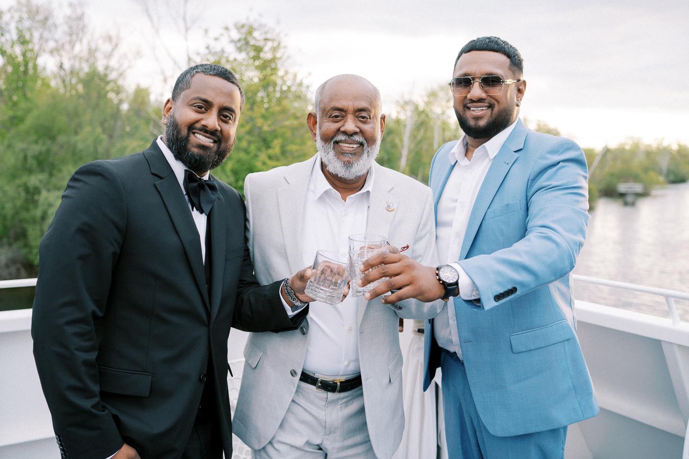 Three men dressed in formal suits, standing on a boat and toasting with clear glasses, surrounded by lush greenery and a serene river in the background.