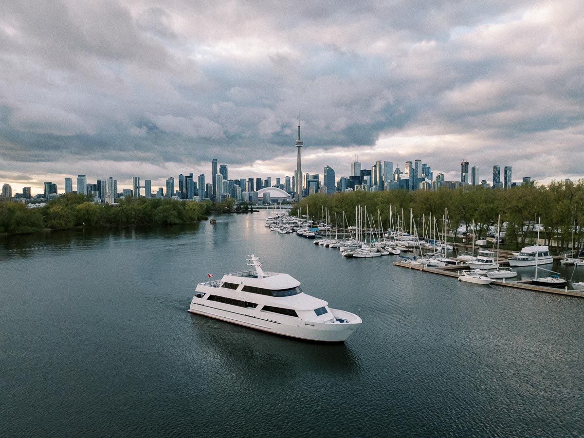 A luxurious white yacht sailing in a calm river with the Toronto skyline and CN Tower in the background under a cloudy sky.