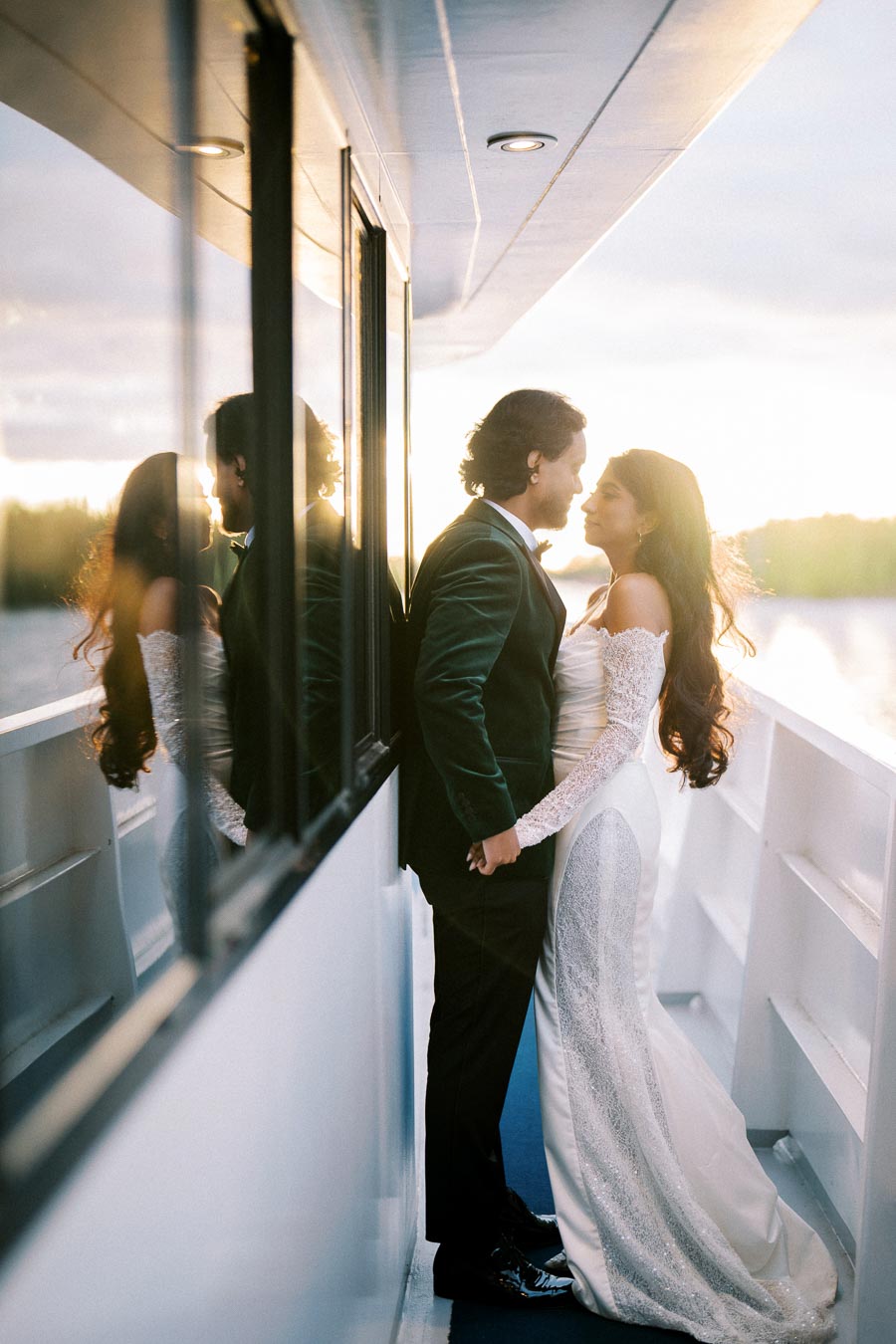 A bride and groom share an intimate moment on a boat at sunset, with the sun casting a warm glow over the water. The bride wears an elegant white gown, and the groom is in a formal suit, holding hands. Reflections of the couple are visible on the boat's windows.