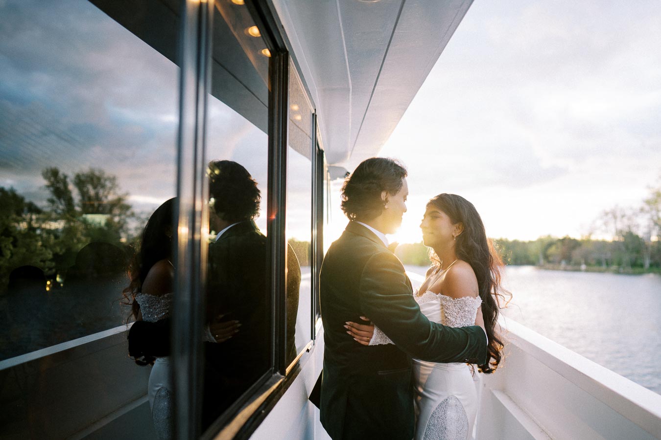 A couple embraced on a boat during sunset, their reflection visible in the window, surrounded by a serene river and trees in the background.