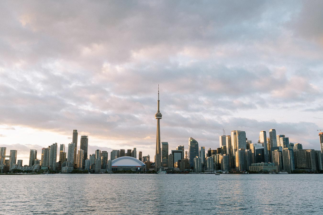 Toronto skyline at sunset featuring the CN Tower and downtown skyscrapers reflecting on Lake Ontario under a partly cloudy sky.