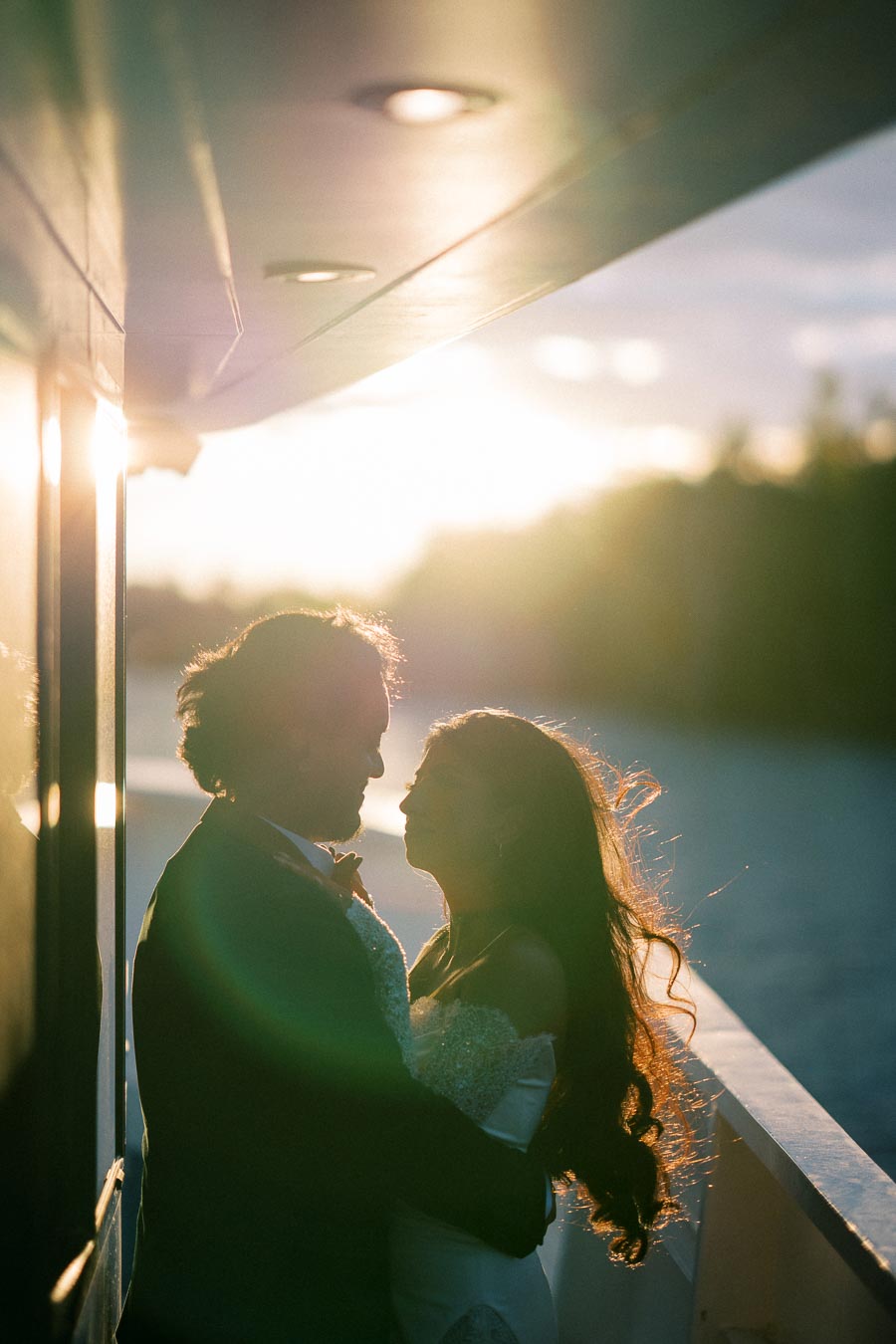 Romantic couple embracing on a boat at sunset, with golden sunlight creating a silhouette effect, highlighting the waterfront backdrop.