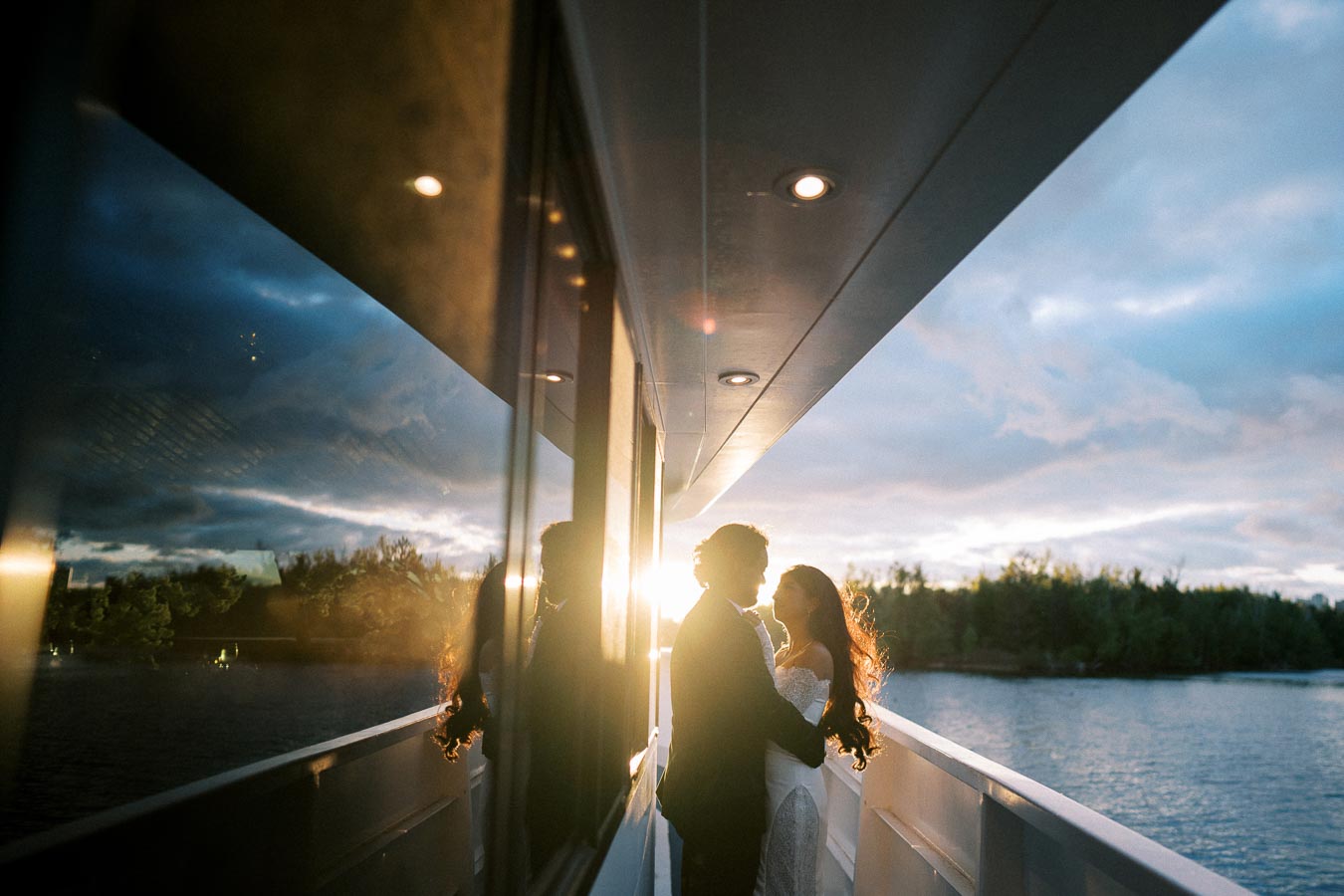 A couple embraces on a boat at sunset, with their silhouettes reflecting on the glass, surrounded by a serene lake and lush greenery.