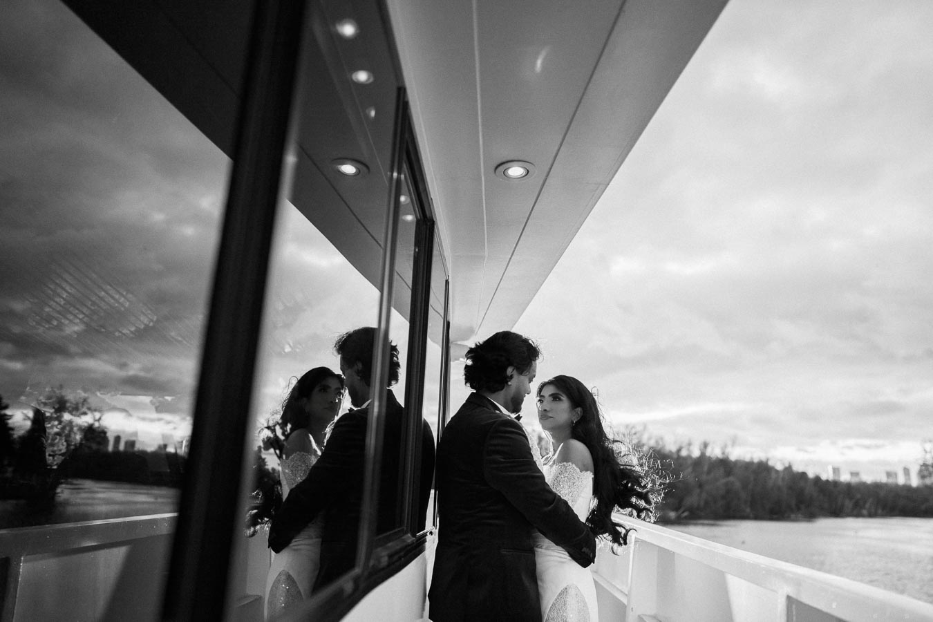 Bride and groom embracing on a boat deck, with reflections in the window, under a cloudy sky.