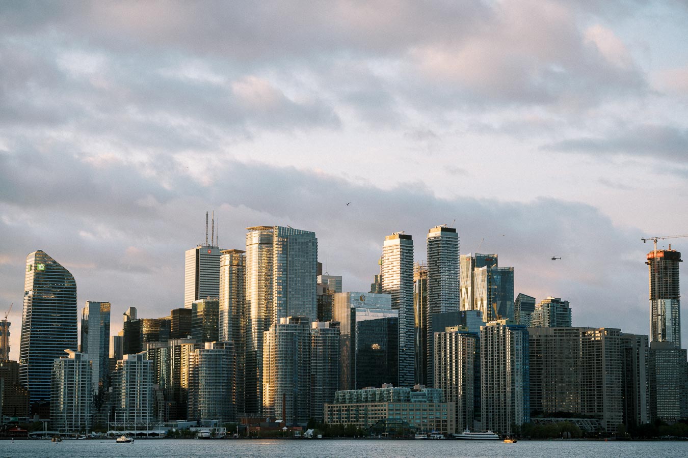 Toronto skyline at sunset featuring modern high-rise buildings and waterfront view, with a partially cloudy sky in the background.