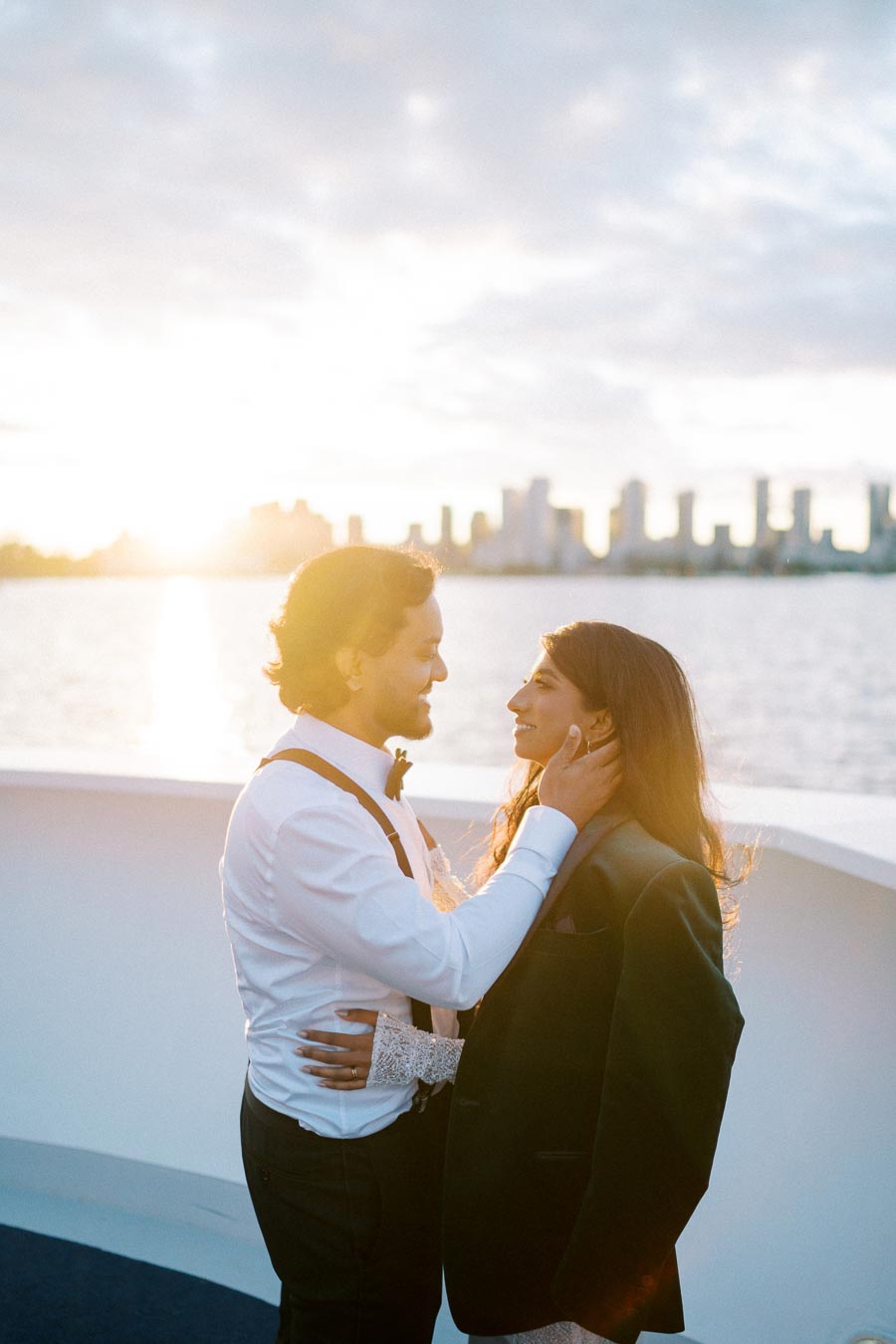 A romantic couple embracing on a boat with the sunset over a city skyline in the background, captured during golden hour.