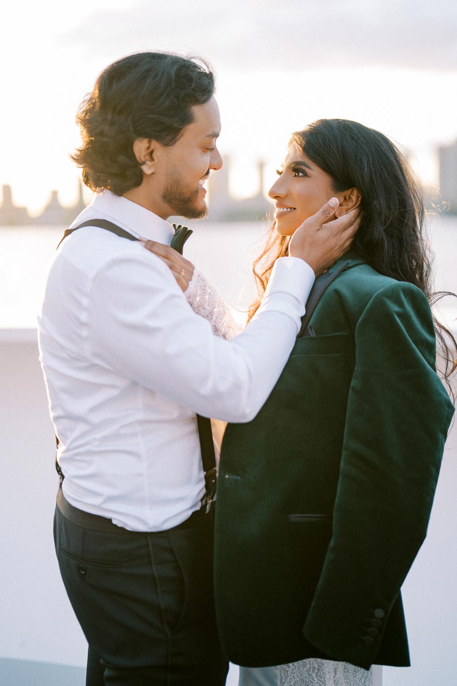 A couple sharing an intimate moment on a rooftop with a city skyline in the background, dressed in stylish formal attire during sunset.