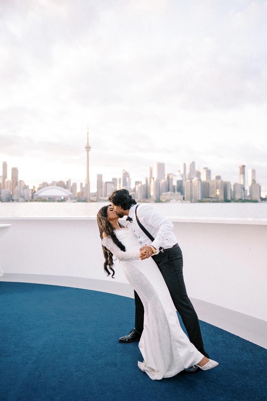 A couple shares a romantic kiss against the backdrop of a city skyline and a tower, possibly the CN Tower in Toronto, with clear skies overhead. The woman is in a white dress and the man in formal attire.