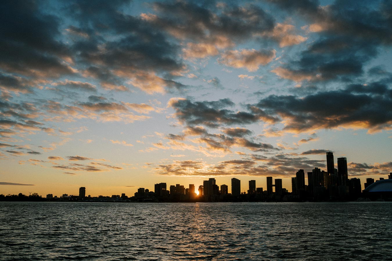 Sunset view of a city skyline with silhouetted skyscrapers against a colorful sky and clouds, overlooking a calm body of water in the foreground.