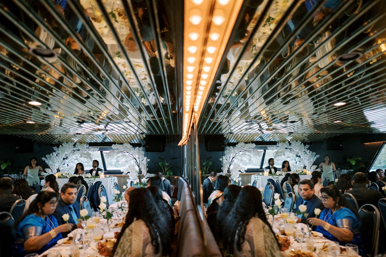 Luxurious dining event aboard a boat with elegantly dressed guests, ornate table settings, and a mirrored ceiling creating a reflective ambiance.