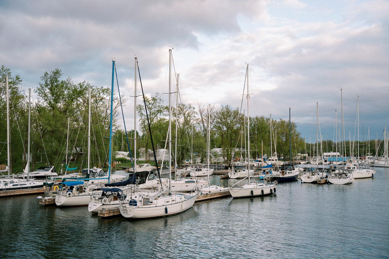 Marina with multiple sailboats docked on calm water, surrounded by lush green trees and a partly cloudy sky.