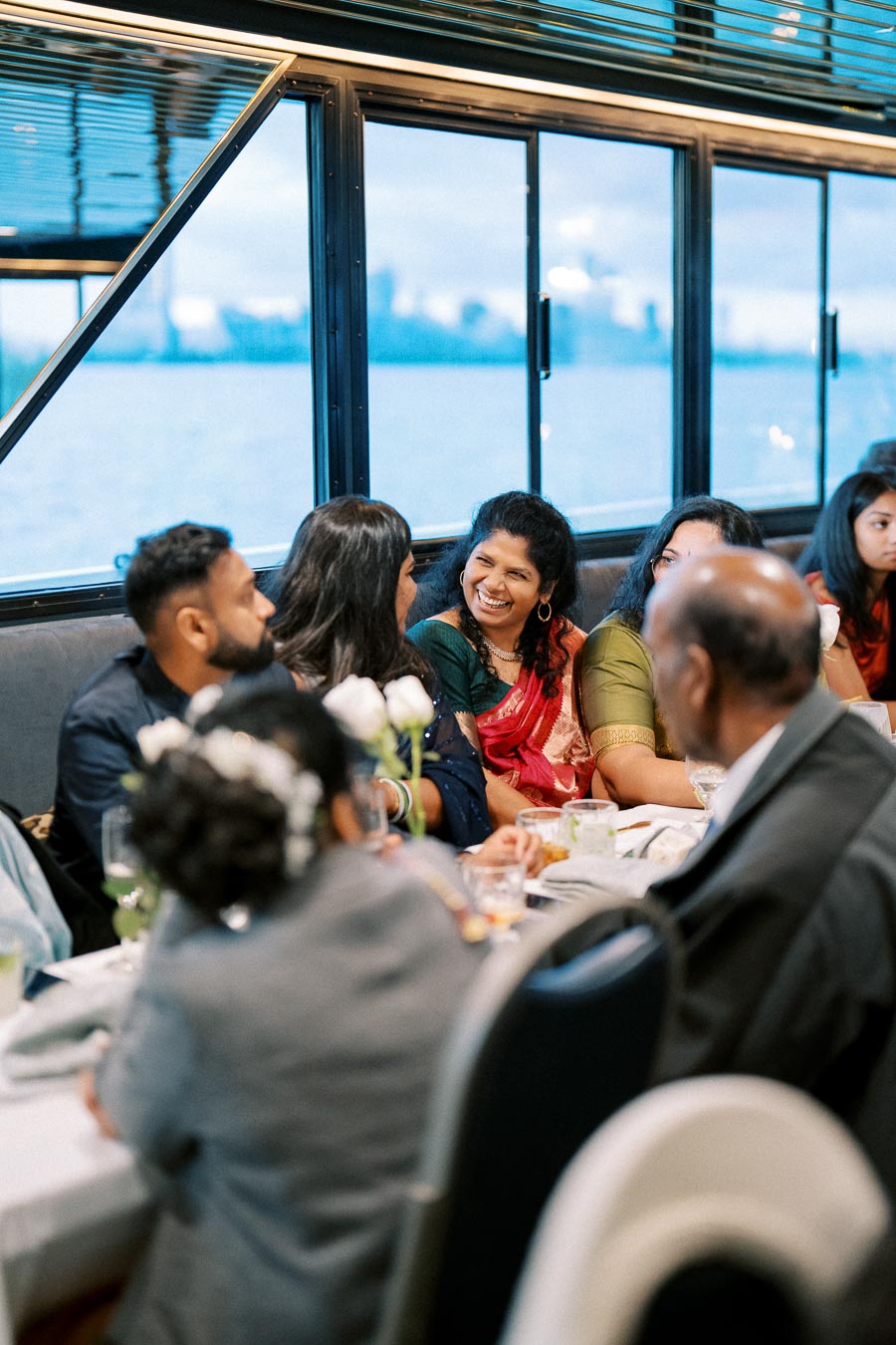 Group of people enjoying a conversation at a formal gathering on a boat, with a scenic view of water and city skyline visible through the windows.