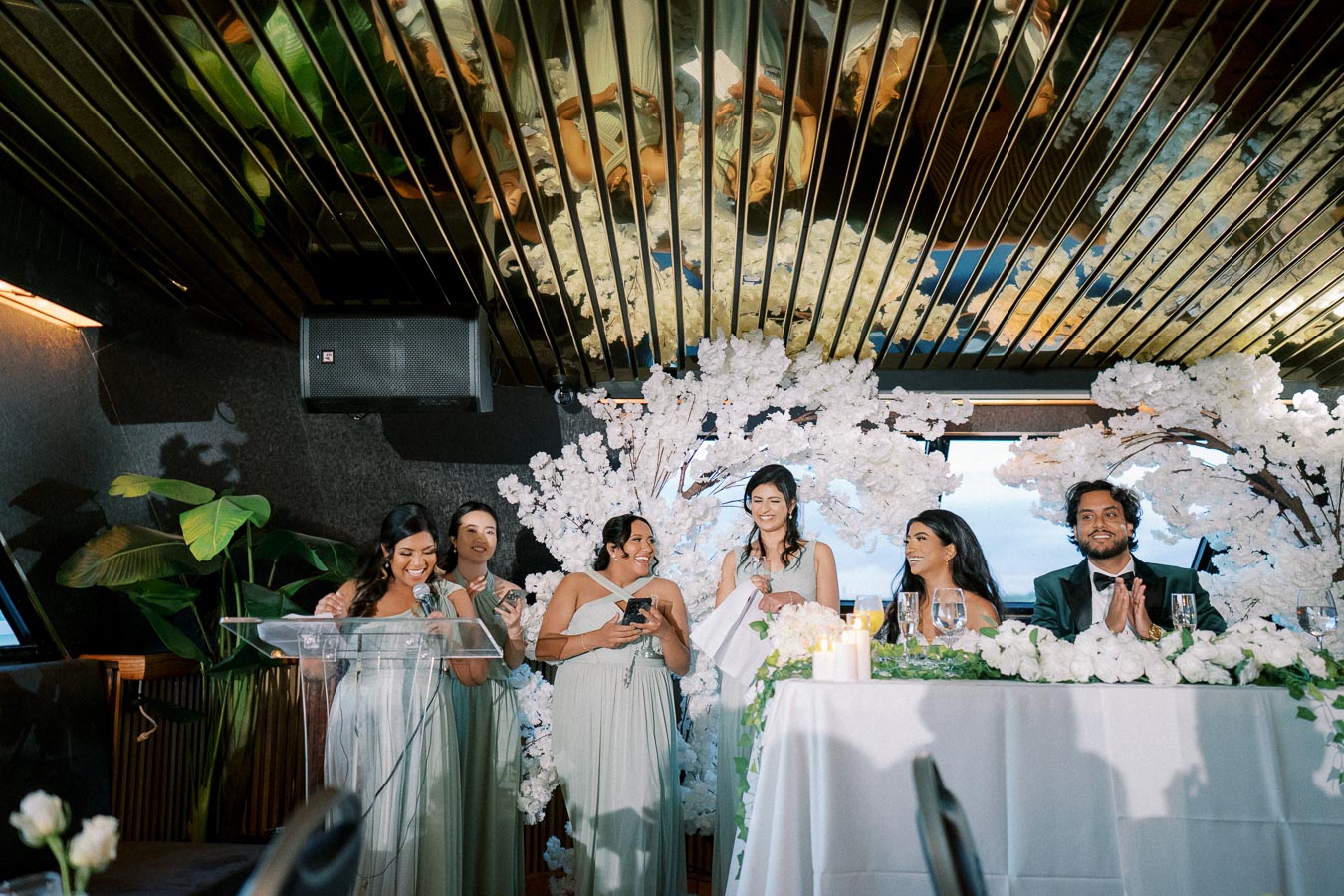 A group of bridesmaids in light green dresses delivering a speech at a wedding reception, standing next to a table adorned with white flowers and greenery, with the bride and groom smiling and clapping in the background.