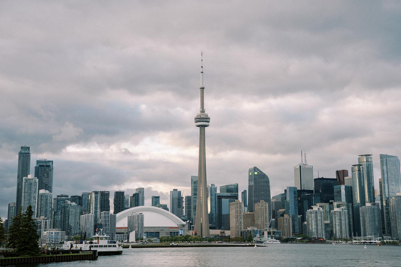 Toronto skyline featuring the CN Tower against a cloudy sky, with urban skyscrapers and waterfront in the foreground.