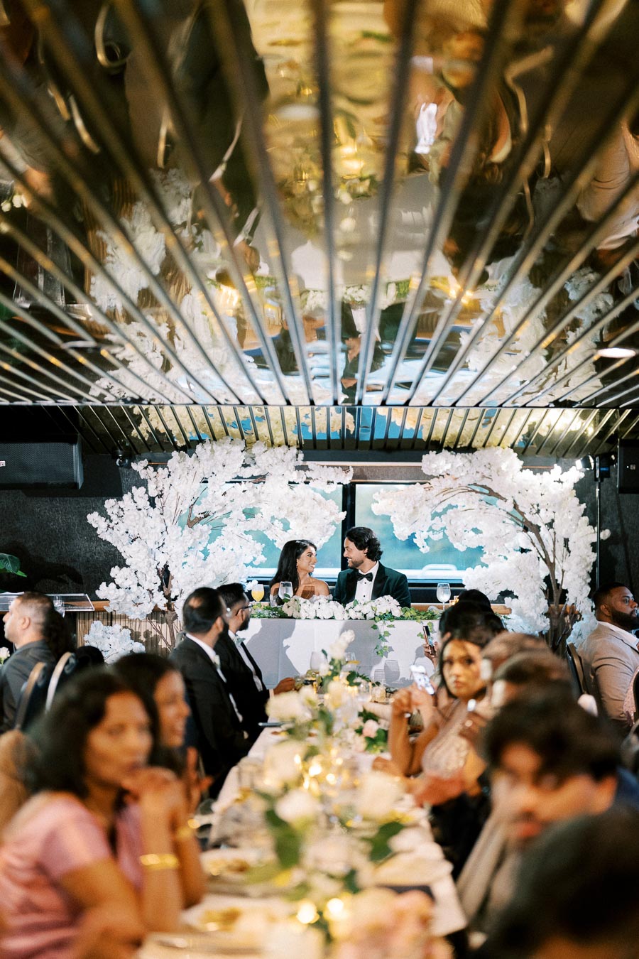 A wedding couple seated at a decorative head table with white floral arrangements, surrounded by guests in formal attire at a reception, reflected in a mirrored ceiling.