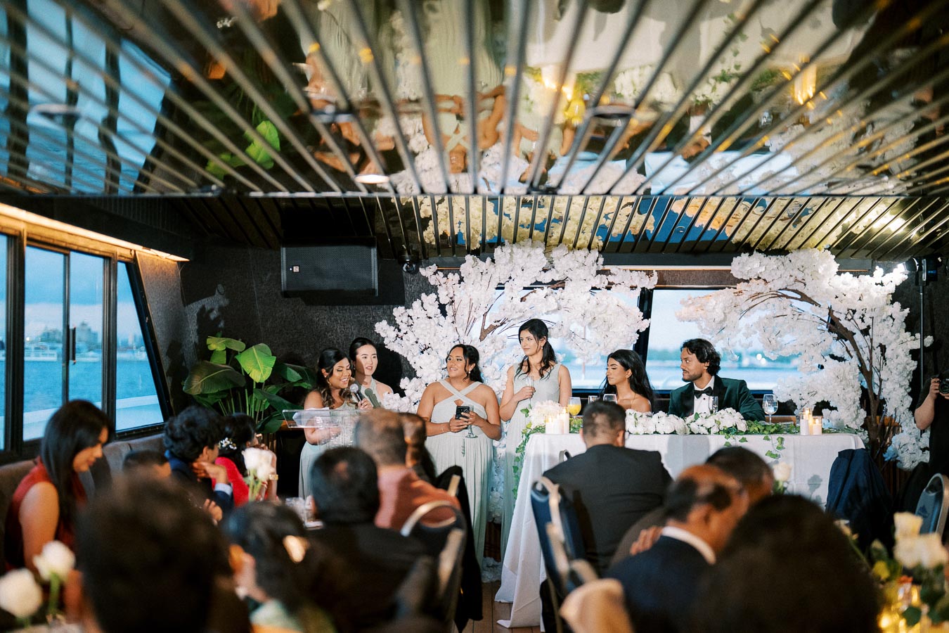 Group of bridesmaids giving a speech at a wedding reception inside a beautifully decorated venue with large windows providing a view of the water. The head table is adorned with floral arrangements and candles, creating an elegant and celebratory atmosphere.
