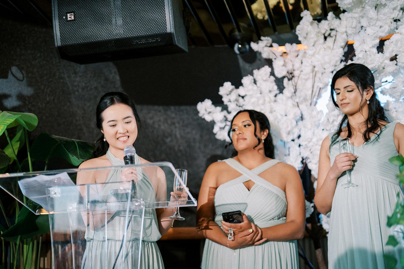 Three women in elegant dresses giving a speech at a wedding reception, holding microphones and champagne glasses, with a floral backdrop.