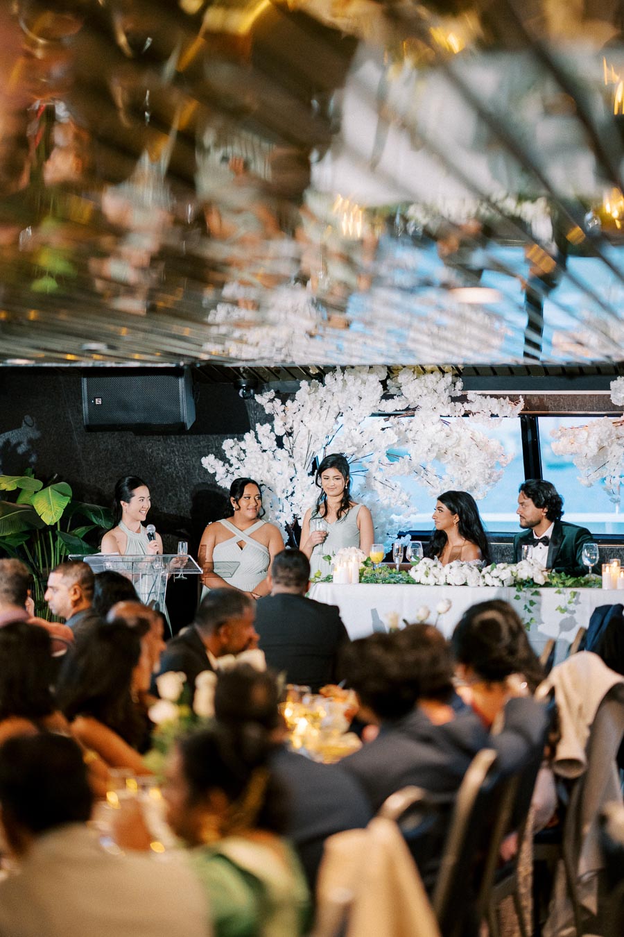 Wedding reception scene with four people speaking at a head table, decorated with white flowers and candles. The audience is seated and appears engaged in a formal setting.