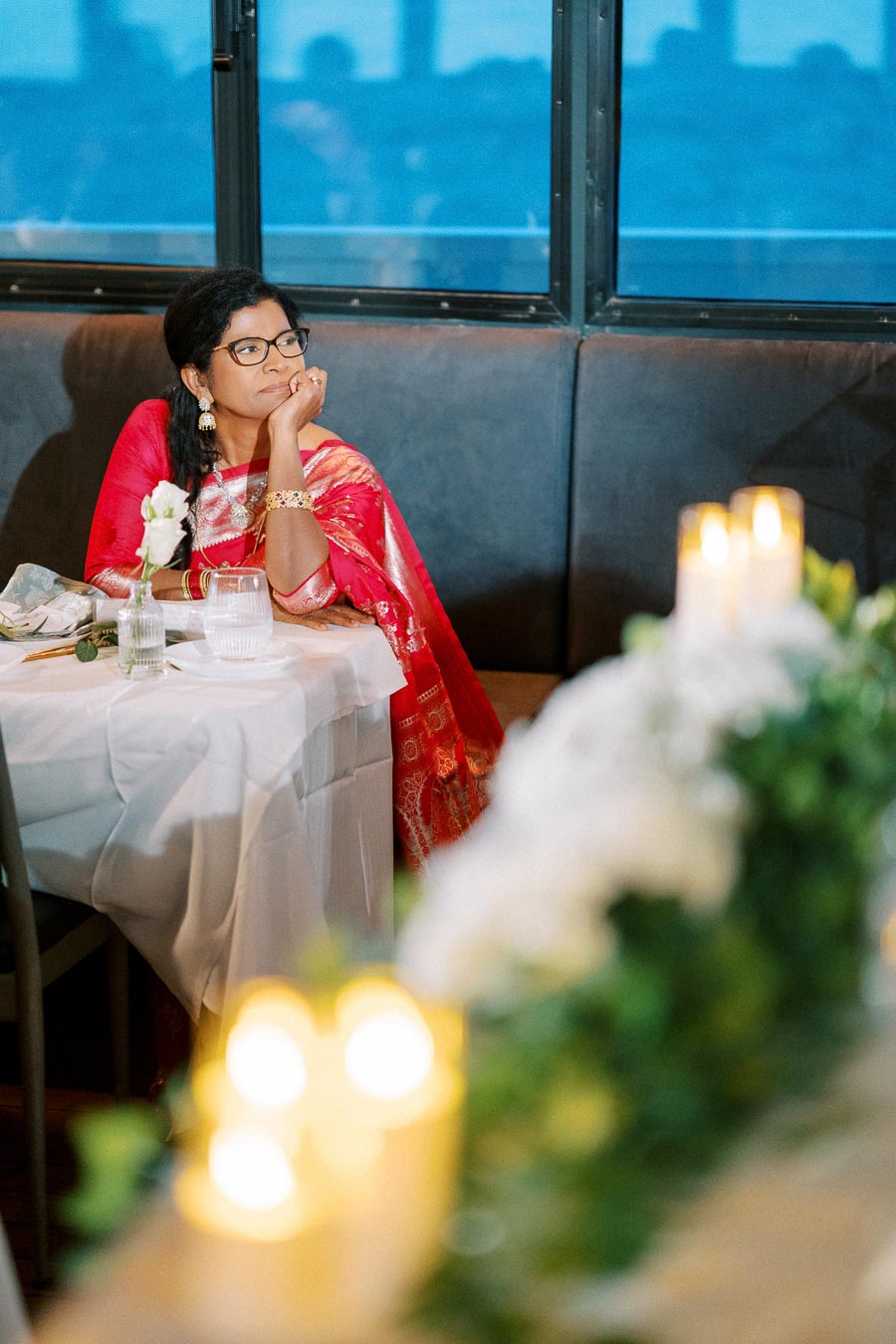 A woman in a red saree sits at a table with a thoughtful expression, surrounded by elegant table settings and candlelight, near a large window.
