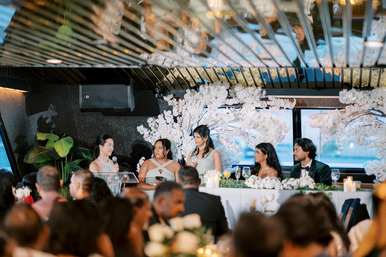 Wedding reception with a woman giving a speech at a beautifully decorated venue. A table adorned with white flowers and candles is attended by elegantly dressed guests seated at the head table, creating a festive and joyful atmosphere.