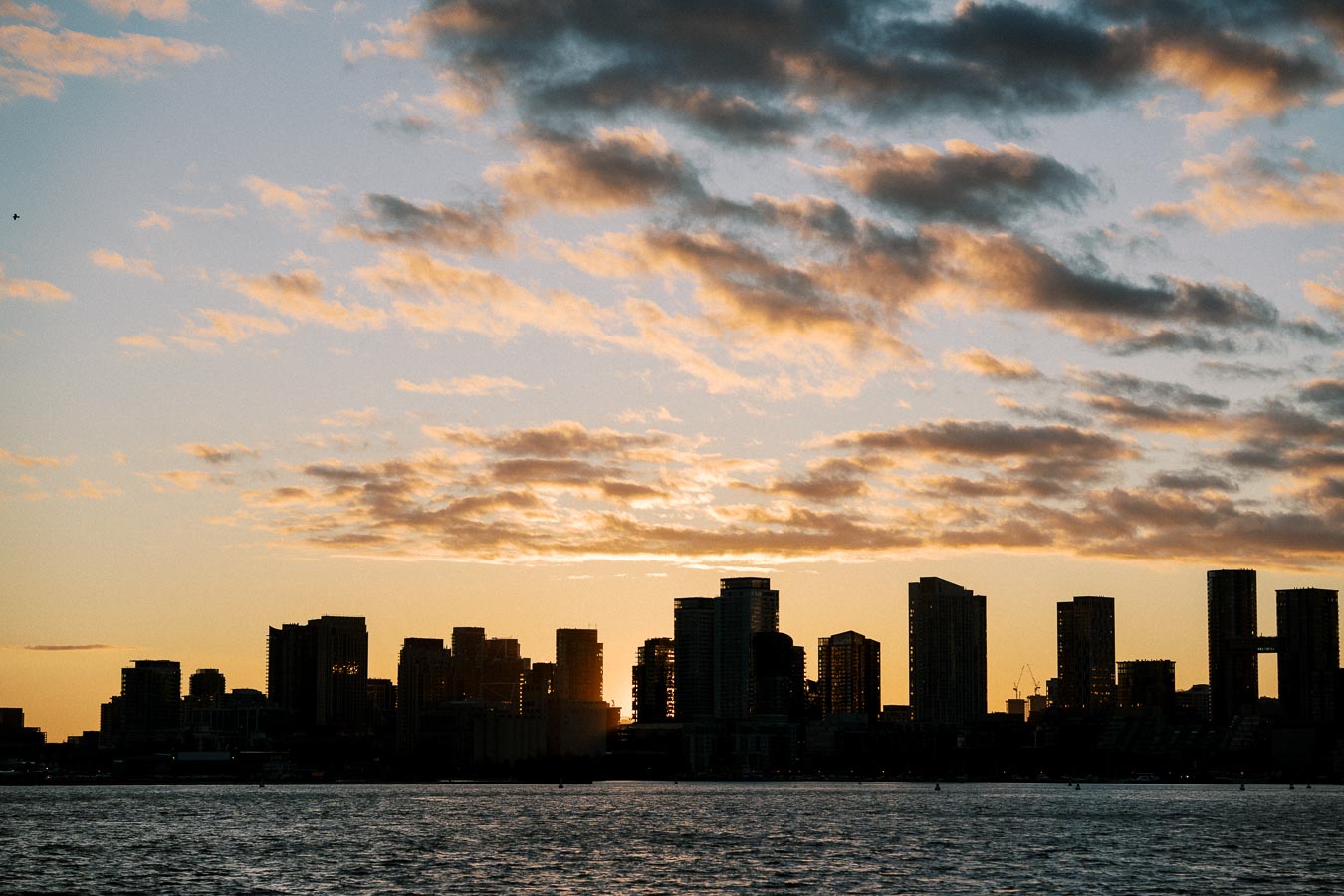 Skyline silhouette of city skyscrapers at sunset, with a partly cloudy sky and a calm body of water in the foreground.