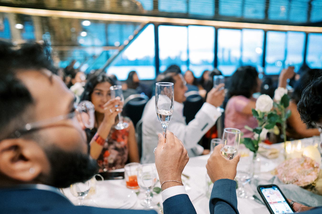 A group of elegantly dressed individuals raising champagne glasses in a celebratory toast at a formal event, with a beautifully decorated table and a panoramic window view in the background.