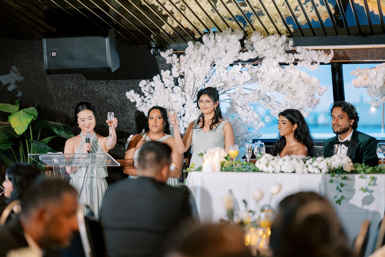 A group of people in formal attire sit at a banquet table adorned with white flowers and greenery. One person stands at a podium, raising a glass for a toast. Behind them is a decorative floral backdrop, creating an elegant wedding reception setting.