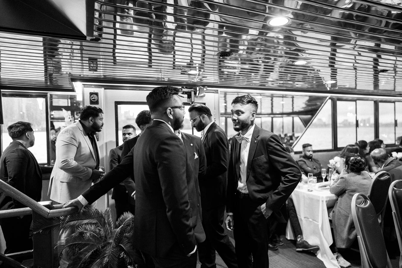A group of men in suits conversing inside an elegantly decorated room with reflective ceilings and large windows, overlooking a scenic waterfront view, during a formal event.