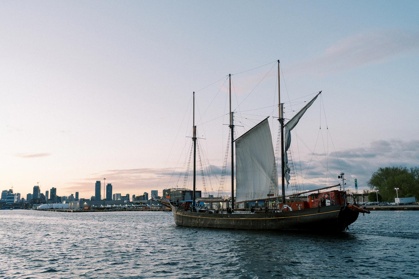 Tall ship sailing near a city skyline at dusk, featuring three masts and partially unfurled sails on a calm urban waterfront.