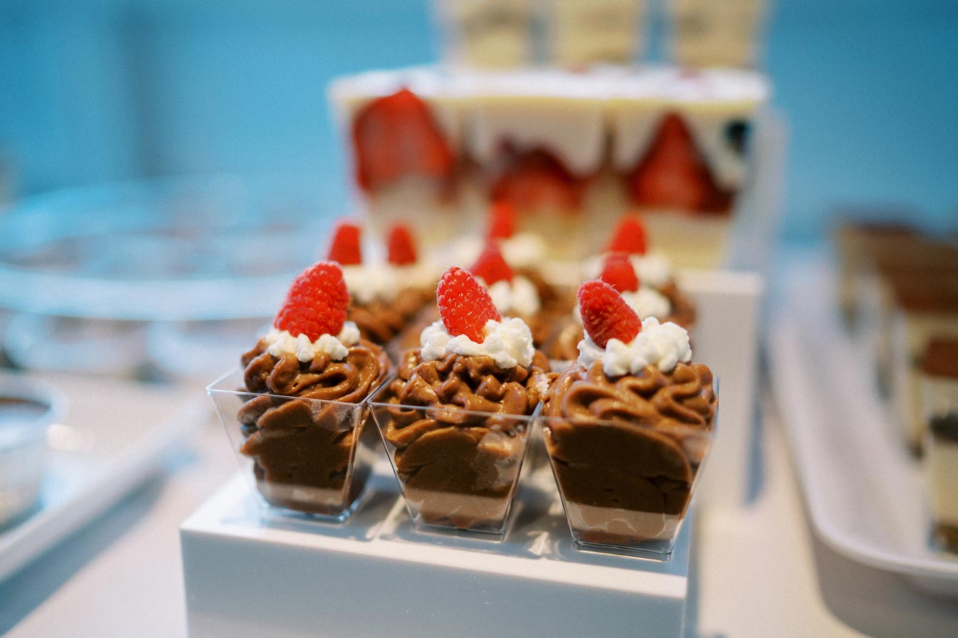 Chocolate mousse desserts served in clear cups, each topped with whipped cream and a fresh raspberry, elegantly displayed on a white platter.