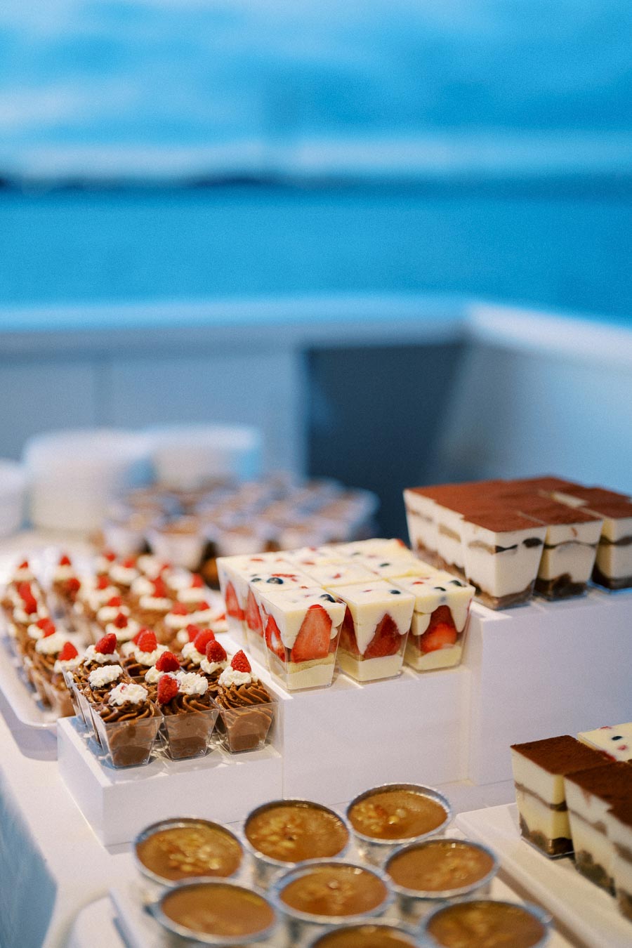 A table adorned with an assortment of elegant desserts, featuring layered fruit parfaits, chocolate mousse with raspberries, and creamy tiramisu cups, set against a serene ocean backdrop.