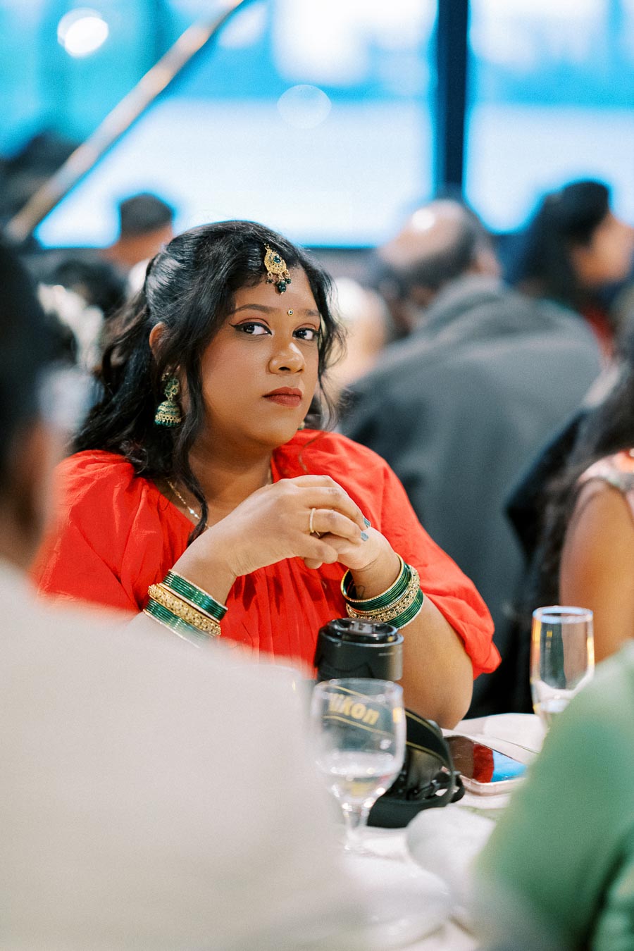 Woman in red traditional attire and jewelry, seated at an event, with a focused expression, surrounded by people and table settings.