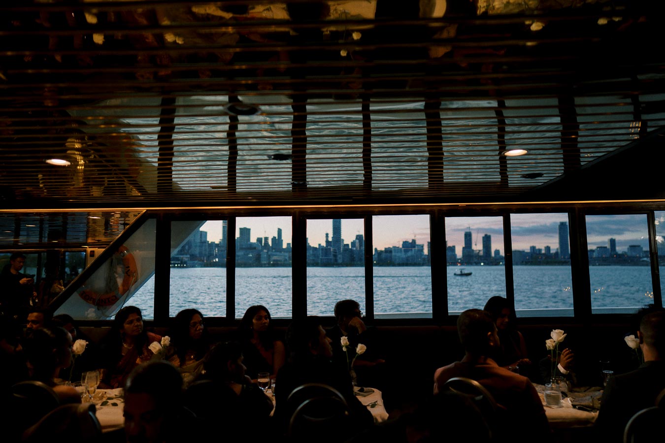 A group of people dining inside a boat or cruise ship with large windows, offering a panoramic view of a city skyline at dusk across a calm river.