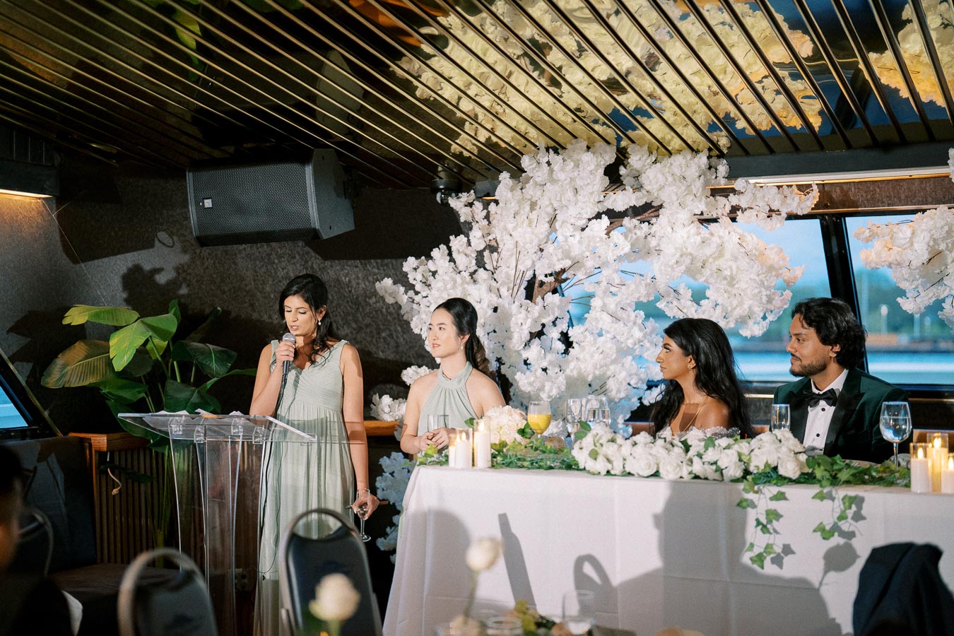 A woman in a light green dress giving a speech at a wedding reception, surrounded by guests seated at a table adorned with white flowers and candles.