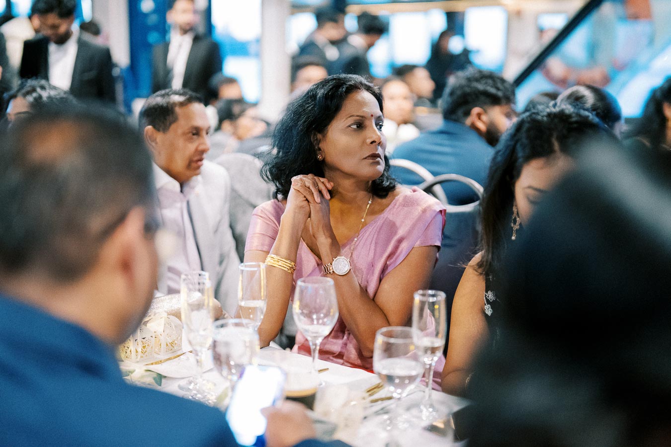 A woman in a pink sari sitting at a formal dining event, surrounded by elegantly dressed people, with wine glasses and plates on the table.