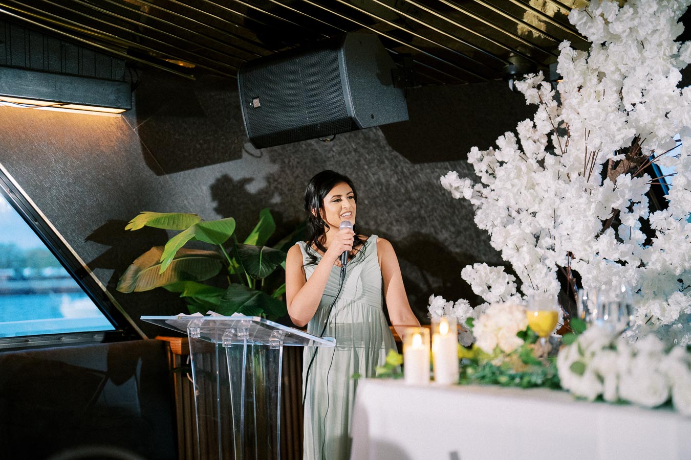 A woman stands at a podium holding a microphone during a formal event, with decorative white flowers and lit candles on the table.