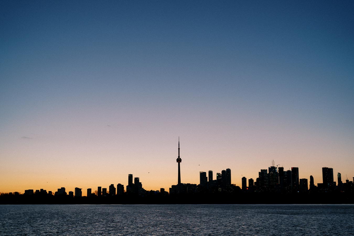 Toronto skyline with CN Tower silhouetted against a colorful sunset, viewed from across a calm body of water.
