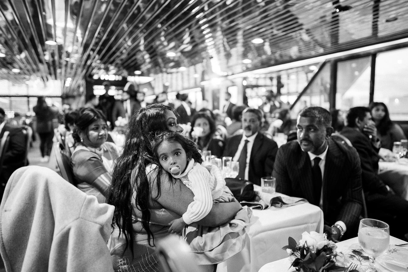 Black and white photo of a family gathering at a restaurant, featuring a woman holding a young child with a pacifier, surrounded by adults in formal attire seated at a table with flower decorations and glasses.
