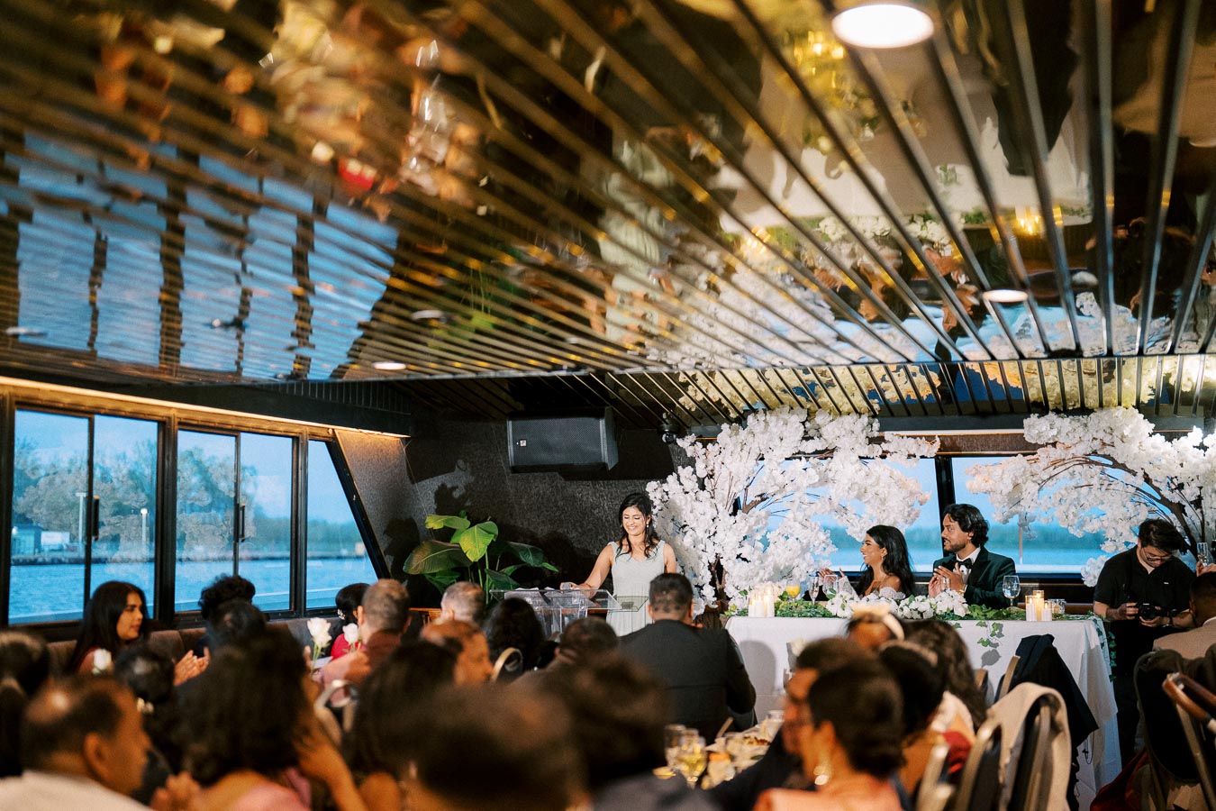 Guests enjoying a formal dinner event on a boat with elegant decorations and floral arrangements, featuring a speaker at the podium and a beautifully set table under ambient lighting.