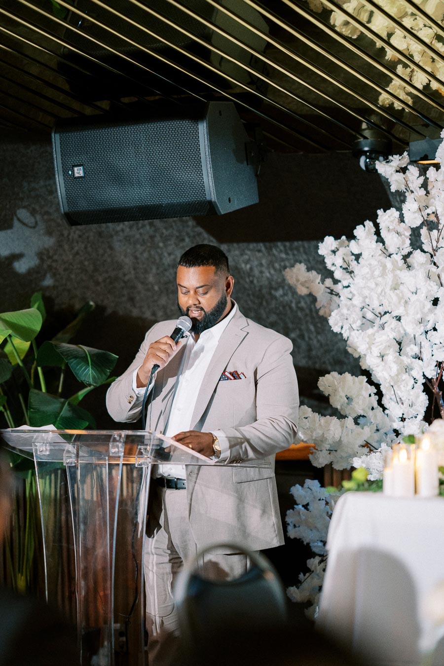 Man in a beige suit speaking into a microphone at a podium, surrounded by elegant floral decorations and ambient lighting, during an indoor event.