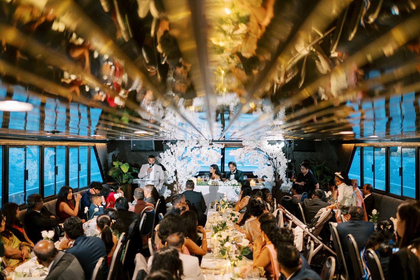 Elegant wedding reception on a scenic boat cruise, featuring a dining area with guests seated at long tables adorned with floral arrangements, a couple at the head table, and a speaker addressing the gathering.