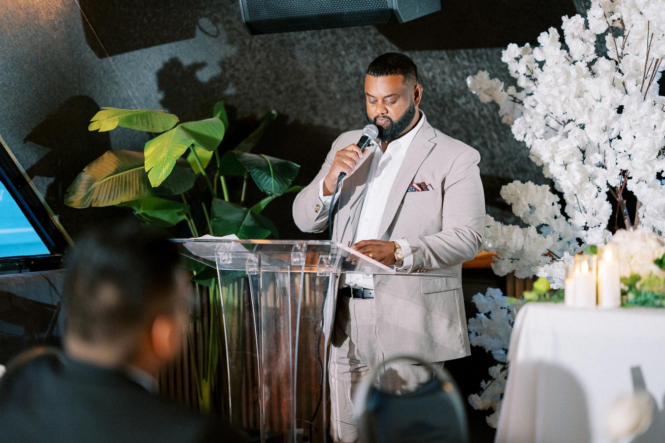 Man in a light suit speaking into a microphone at a clear podium, surrounded by decorative plants and floral arrangements, during an indoor event.