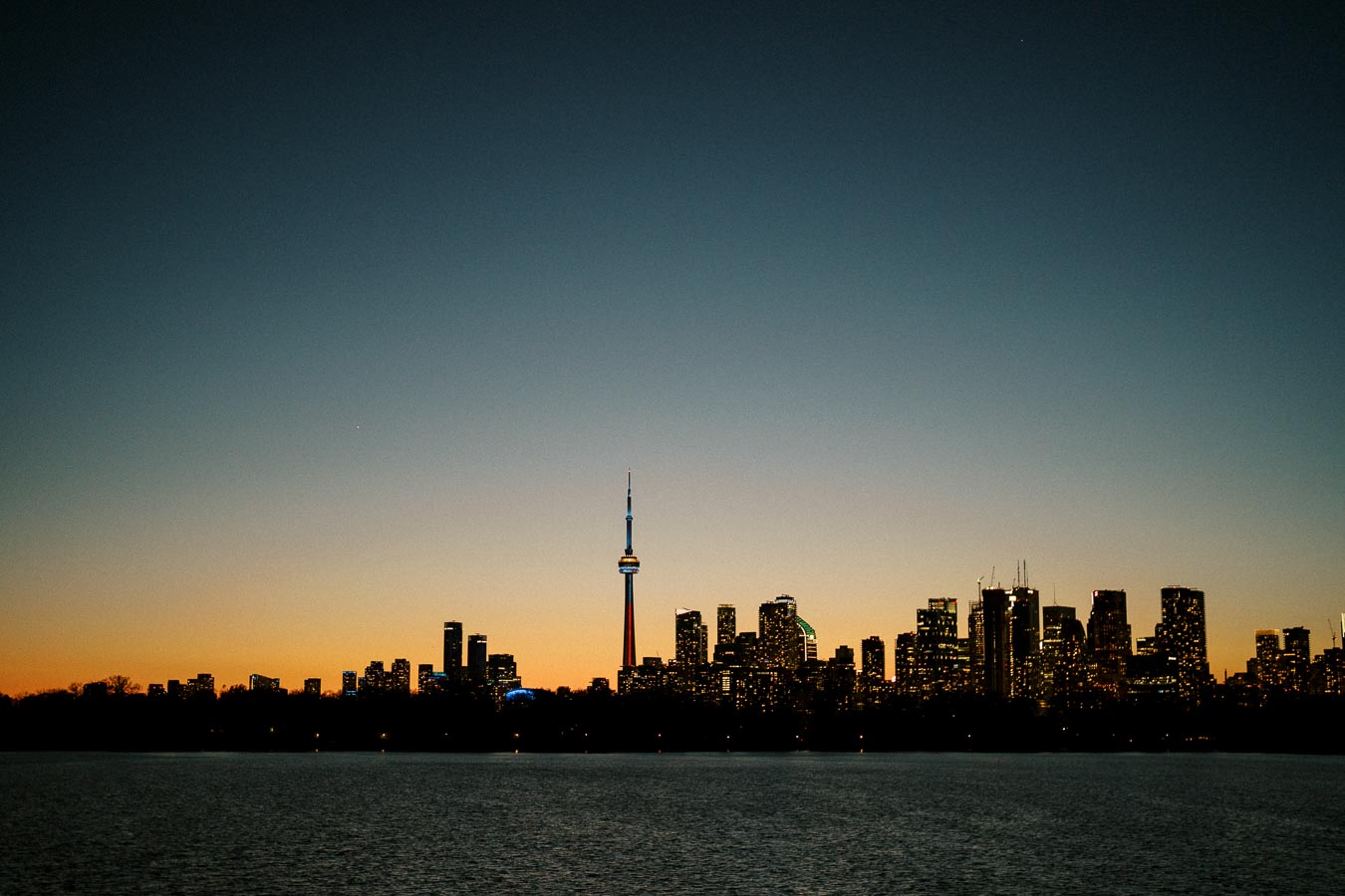 Skyline of a city with a prominent tower silhouetted against a vivid sunset, reflecting on a calm body of water, creating a serene cityscape.