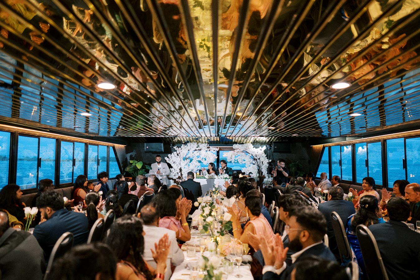 Wedding reception on a boat with guests seated at elegantly decorated tables, featuring floral arrangements and a mirrored ceiling.