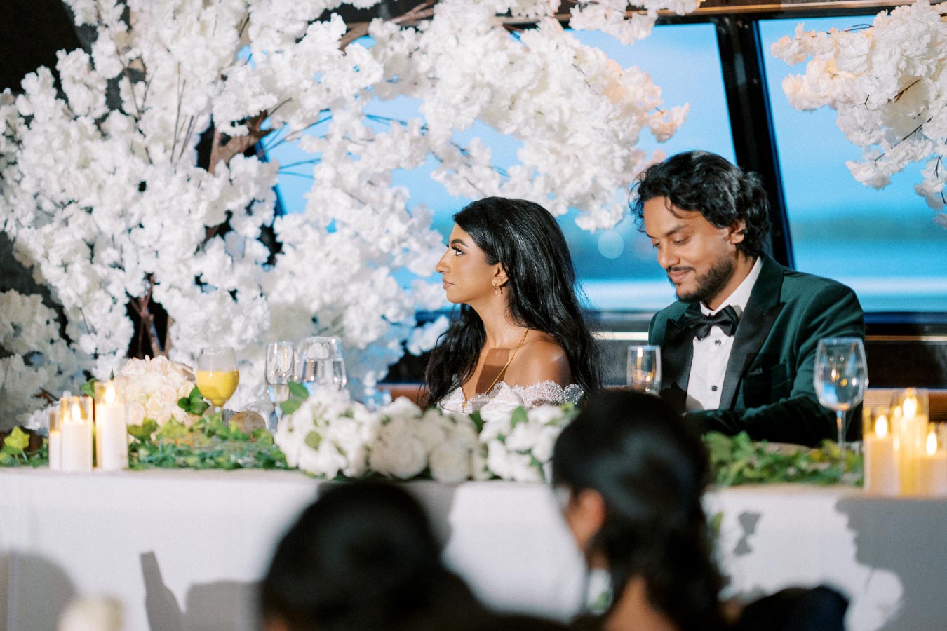 A bride and groom sit at a beautifully decorated wedding table adorned with white flowers and candles, surrounded by elegant table settings and lush floral arrangements, with a picturesque view in the background.