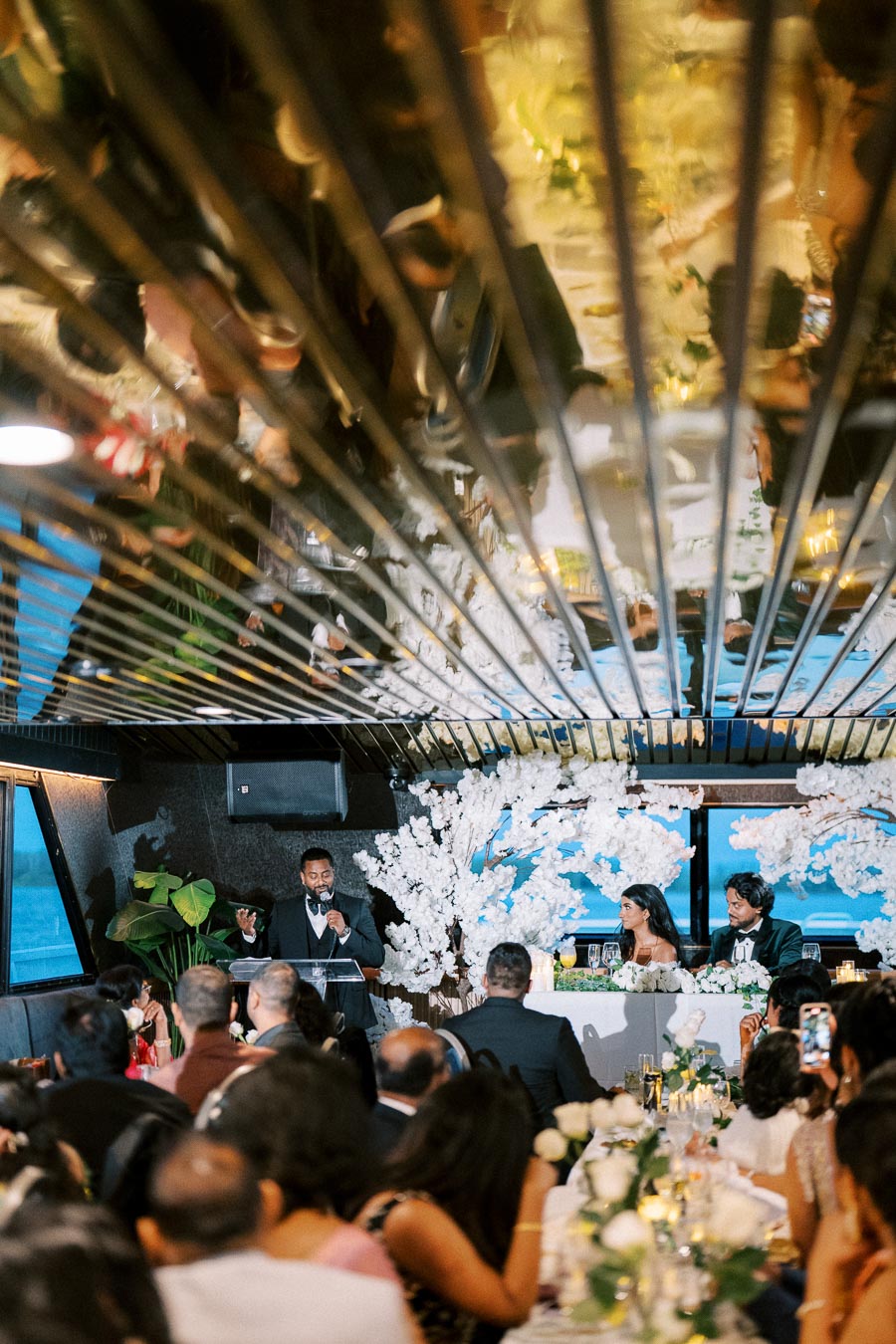 A speaker giving a toast at a wedding reception on a boat, with guests seated around elegantly decorated tables, featuring floral arrangements and a mirrored ceiling.