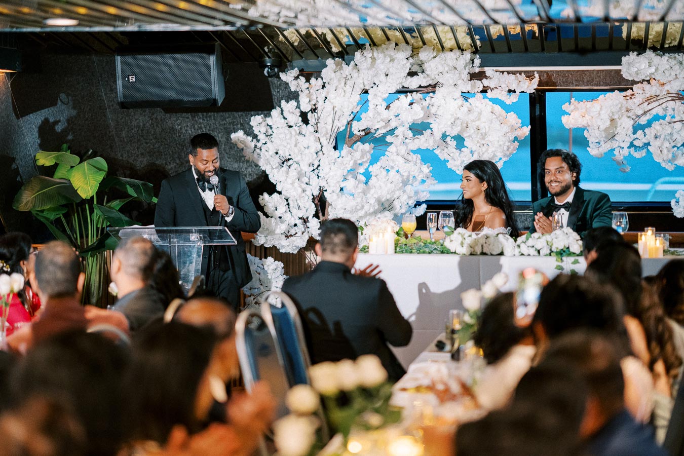 A wedding reception scene with a man giving a speech at a podium, surrounded by elegant white floral decorations. A bride and groom are seated at a table, smiling and clapping, with guests seated in the foreground. The atmosphere is festive and celebratory.