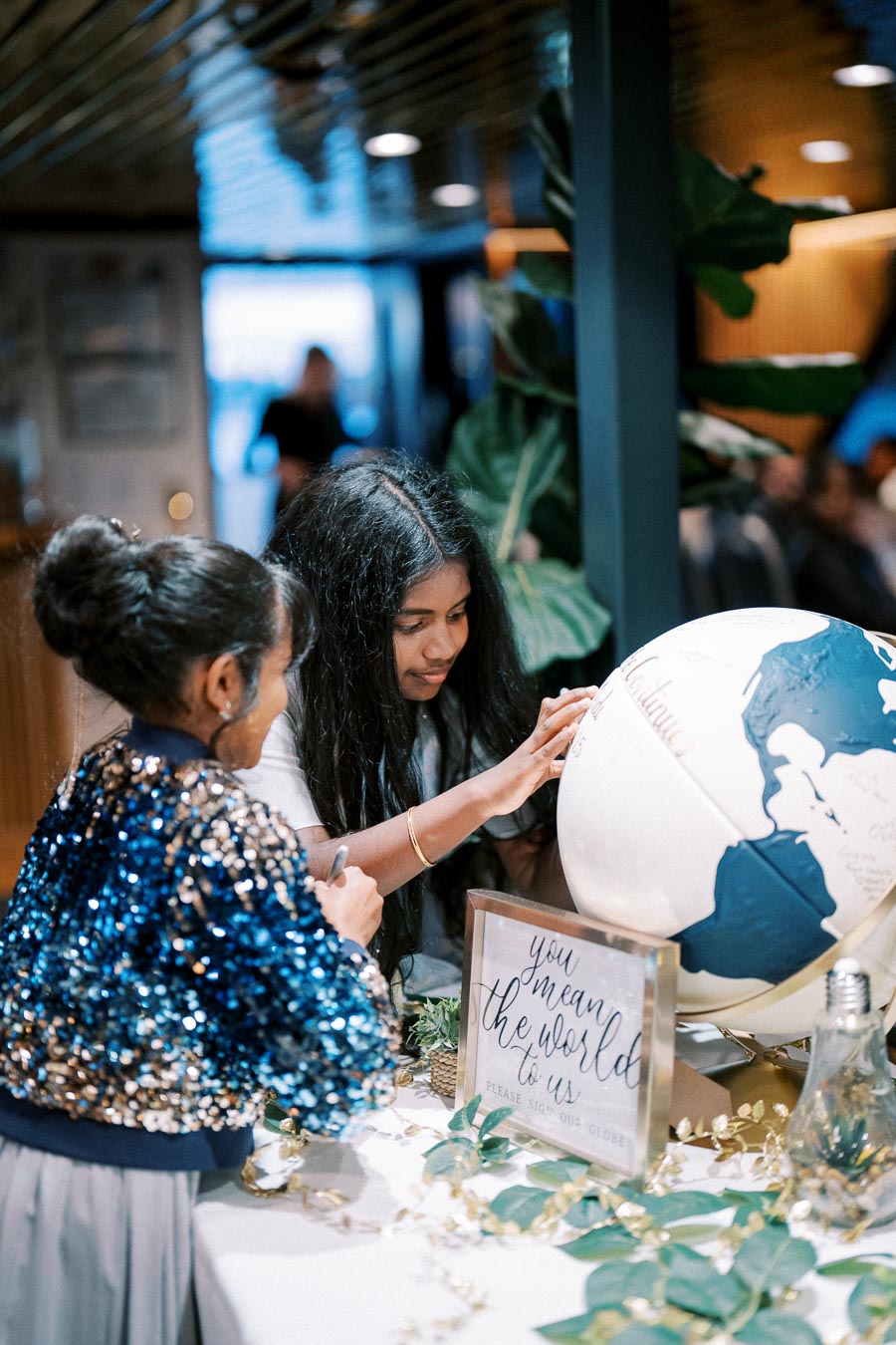 Two people signing a decorative globe at an event, surrounded by greenery and a sign reading “You mean the world to us.”